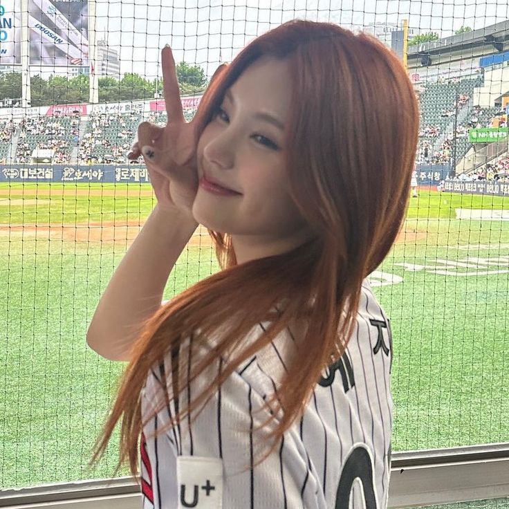 a woman with long red hair standing in front of a baseball field holding her finger up