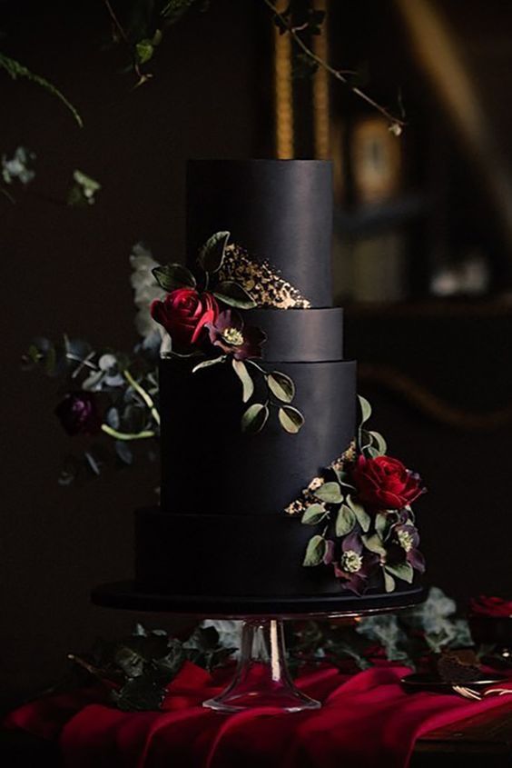 a three tiered black cake with red flowers and leaves on the top, surrounded by greenery