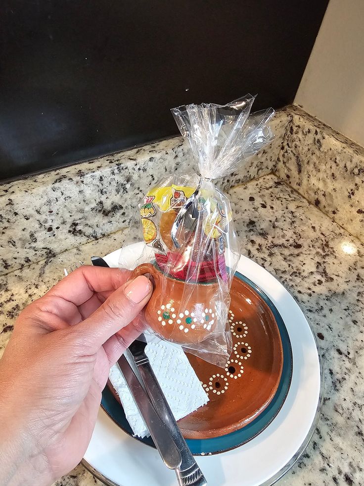 a person holding a knife and fork over a plate with chocolate cake on it, wrapped in cellophane