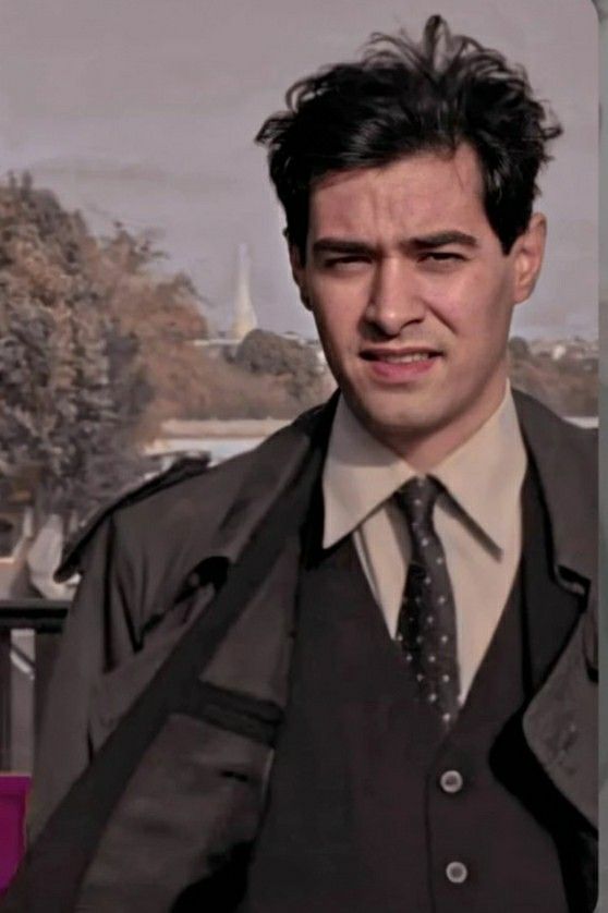 a man wearing a suit and tie standing in front of a fence with trees behind him