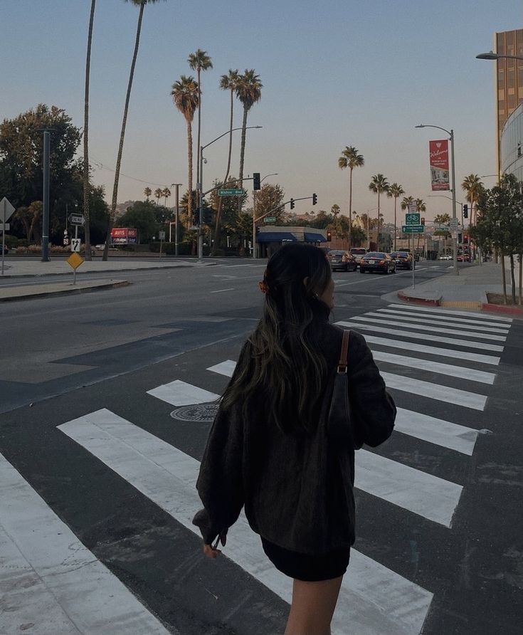 a woman walking across a cross walk in the middle of a street with palm trees