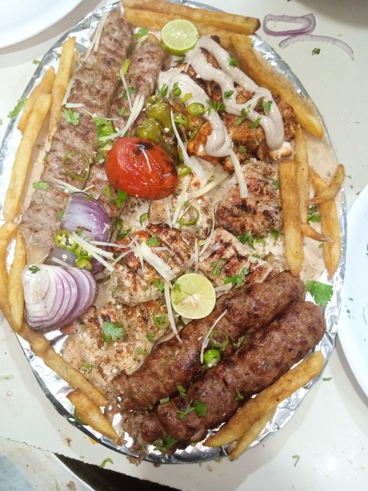 a platter filled with different types of meats and vegetables on tin foil next to french fries