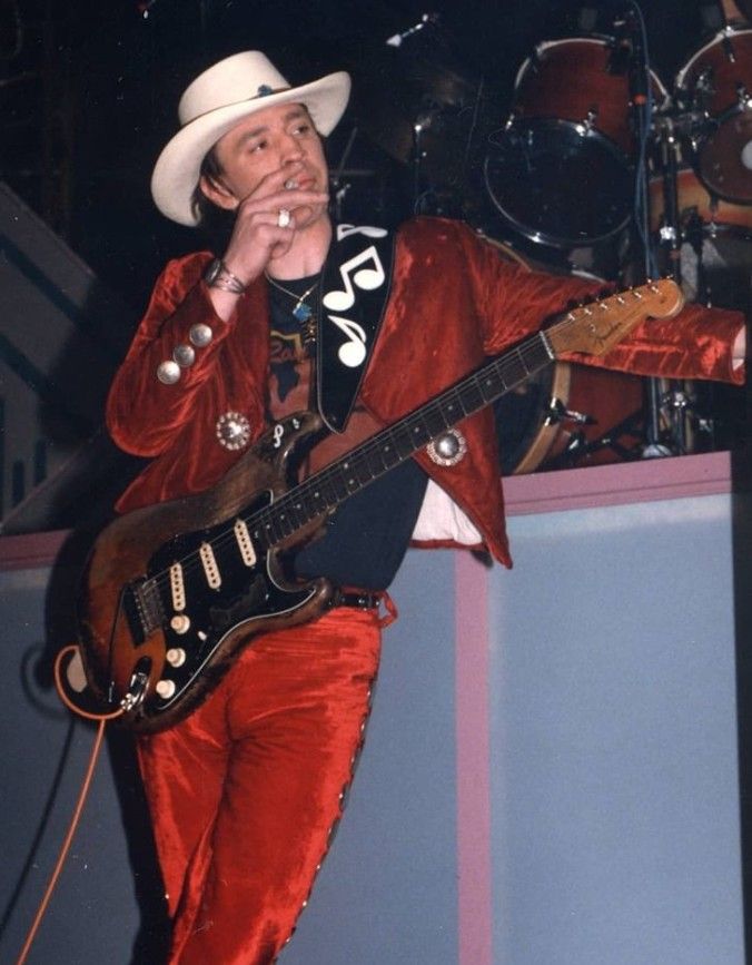 a man in red pants and a white hat is playing the guitar on stage with his hand up to his mouth