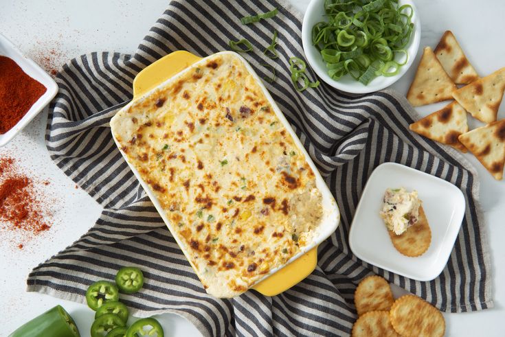 a casserole dish with cheese and other ingredients on a striped towel next to bowls of chili, green peppers, and pita chips