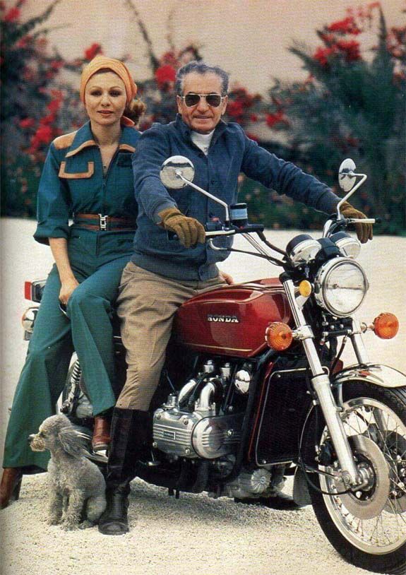 an older man and woman riding on the back of a red motorcycle with flowers in the background