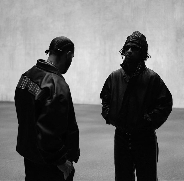 two young men standing next to each other in front of a concrete wall, one with dreadlocks on his head