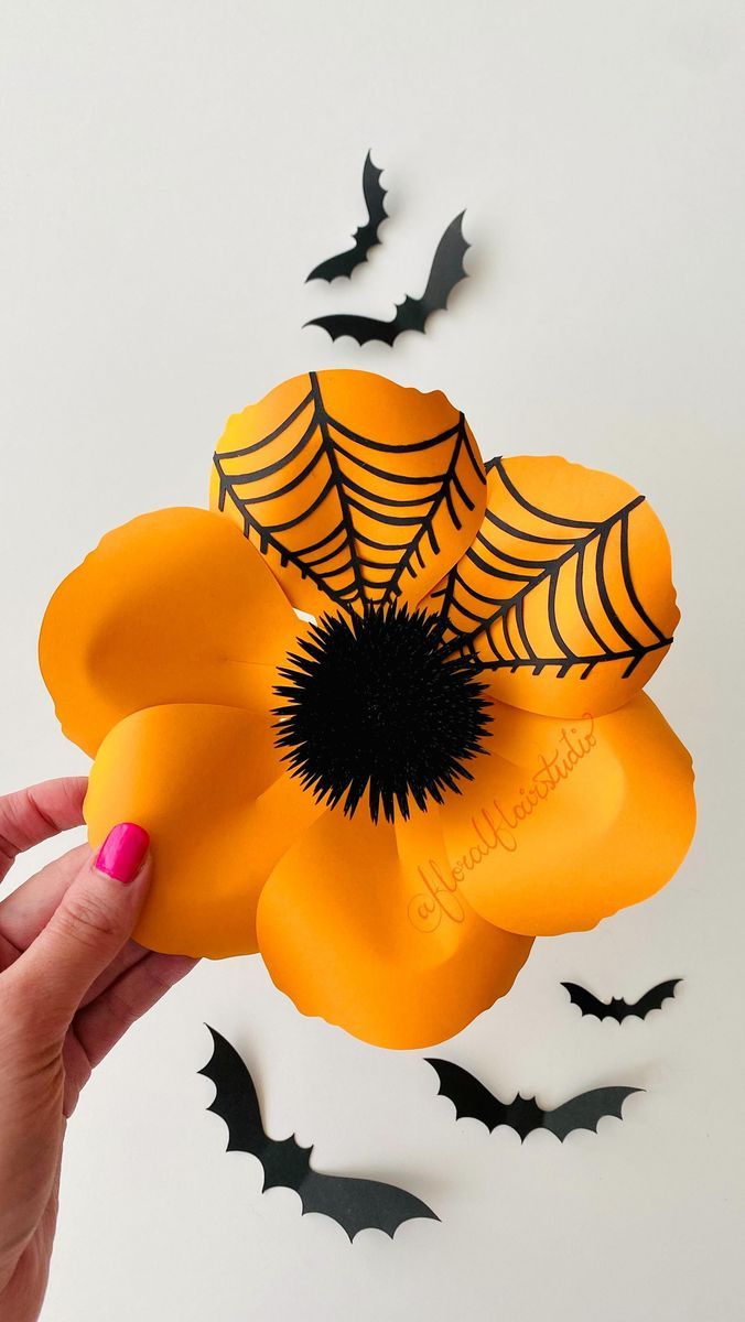 a hand holding an orange paper flower with black spider webs on it and bats in the background