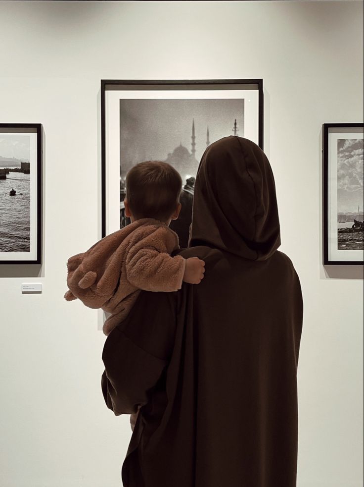 a woman holding a child in her arms while looking at pictures on the wall behind them