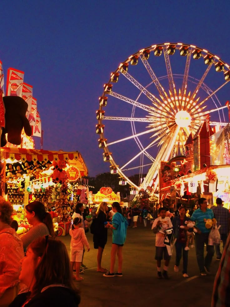 an amusement park at night with people walking around and ferris wheel in the background,