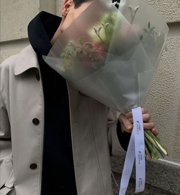 a man holding a bouquet of flowers in front of his face while wearing a trench coat