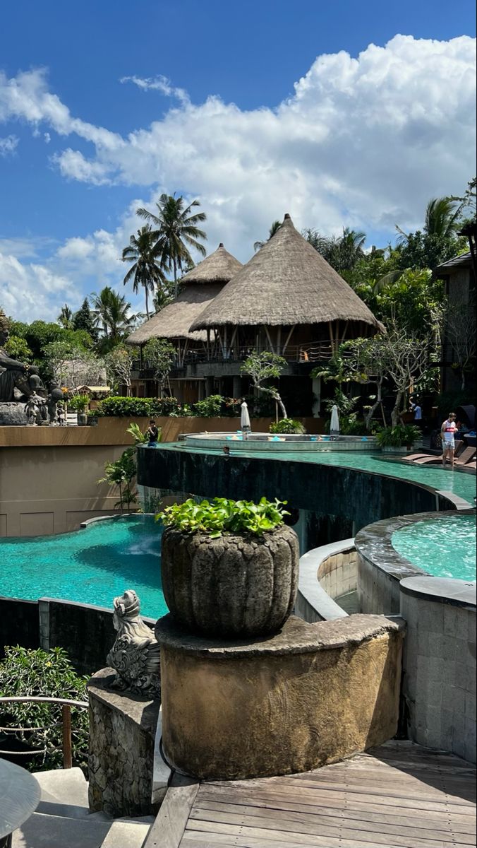 an outdoor swimming pool surrounded by palm trees and thatched roof huts with plants in the middle