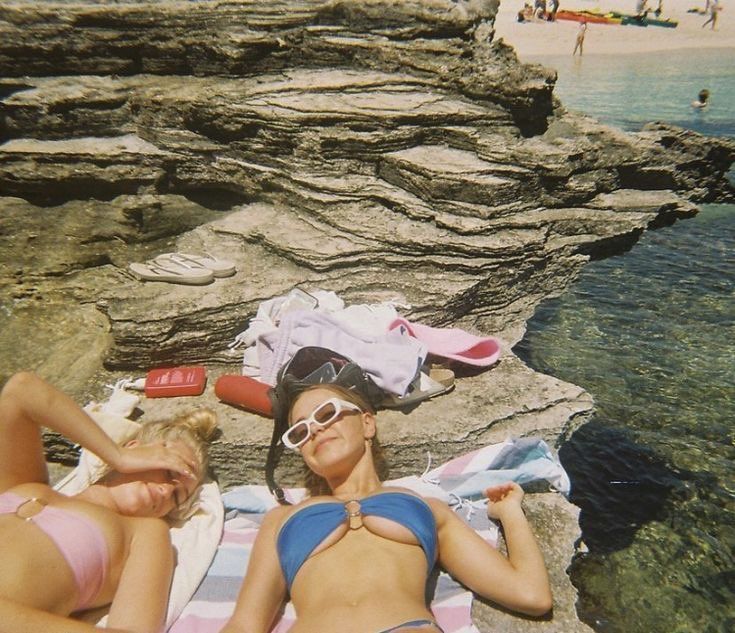 two women in bikinis laying on the rocks by the ocean with towels and flip flops