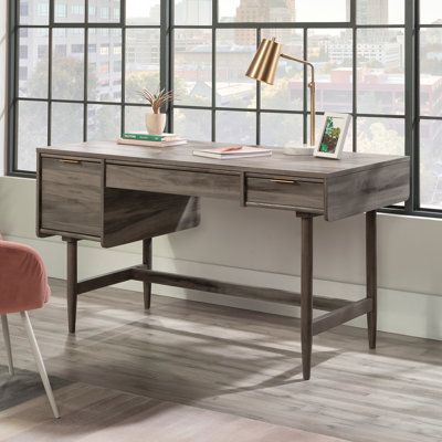 a wooden desk sitting in front of a window next to a chair and table with a lamp on it