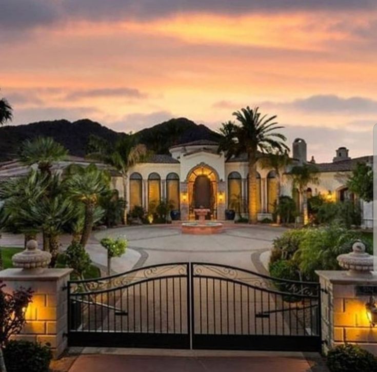 a gated driveway leading to a large home with palm trees and lights on it