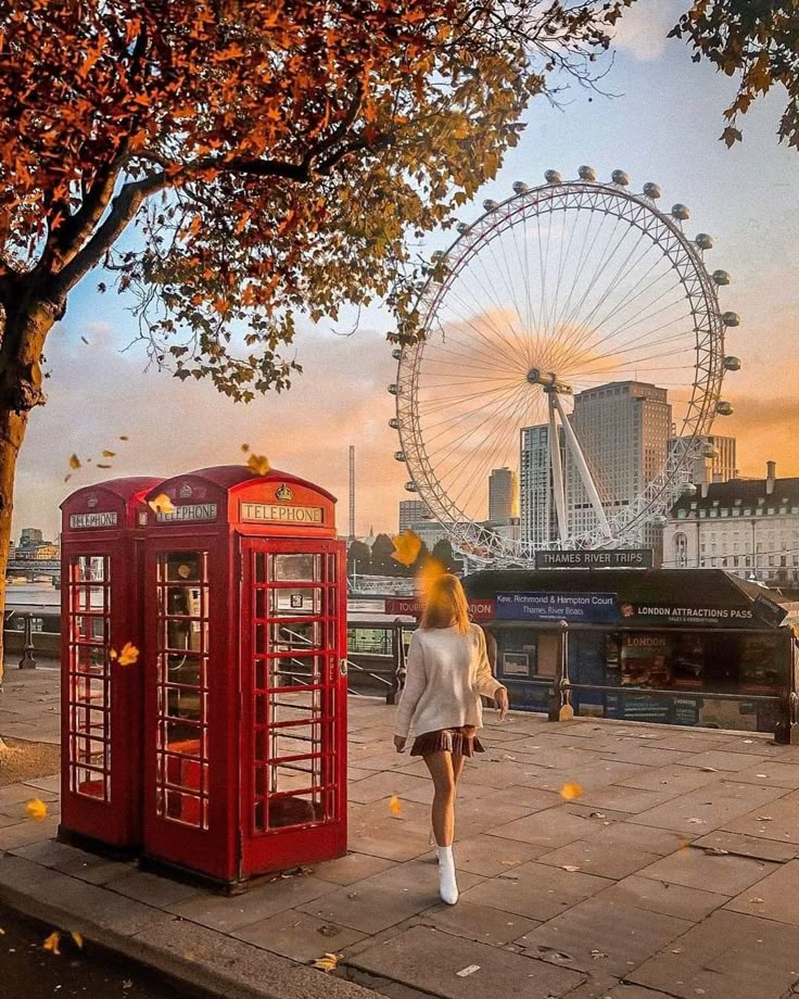 a woman standing next to a red phone booth in front of a ferris wheel at sunset