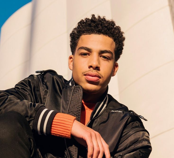 a young man wearing an orange wrist band and black jacket leaning against a white building