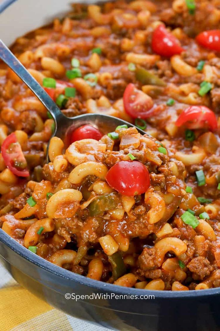 a spoon full of chili macaroni and cheese with tomatoes on top, in a blue casserole dish