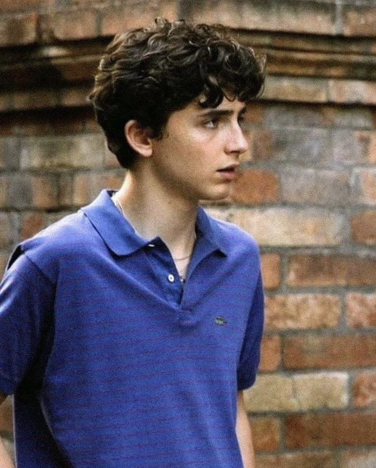 a young man standing in front of a brick wall wearing a blue shirt and jeans