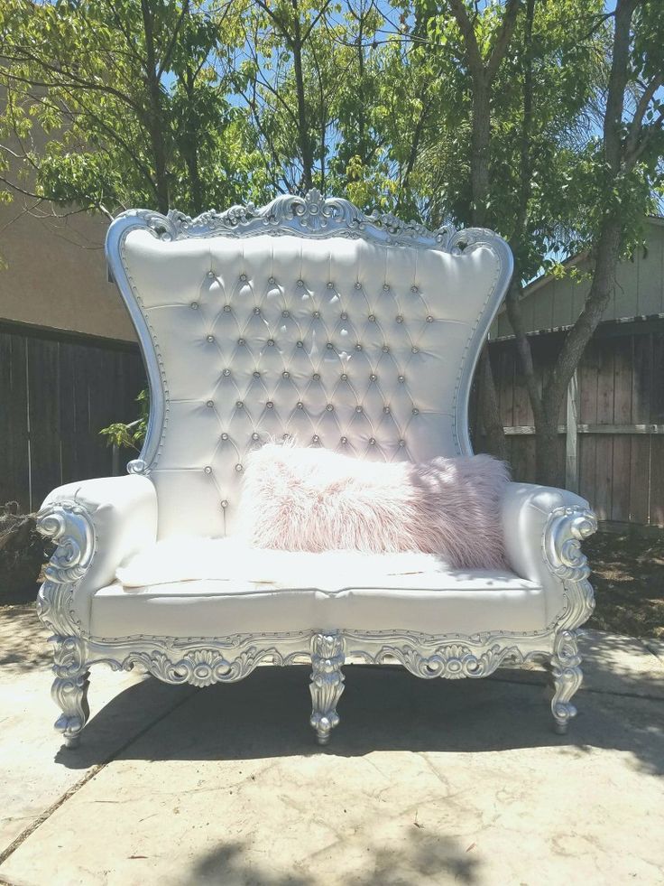 an ornate white chair with pink fur pillow on it's back and legs, in front of a tree