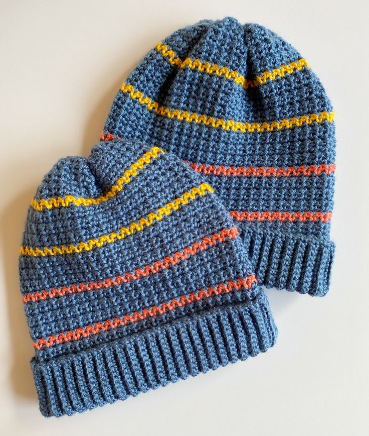 two blue knitted hats sitting next to each other on a white surface with yellow and red stripes