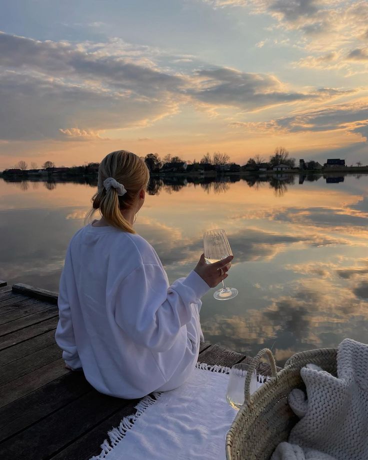 a woman sitting on a dock holding a glass of wine and looking at the water