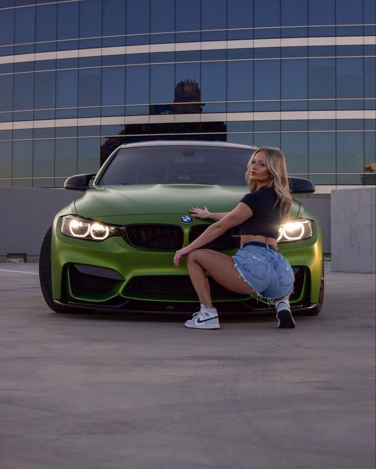 a woman sitting on the hood of a green car in front of a tall building