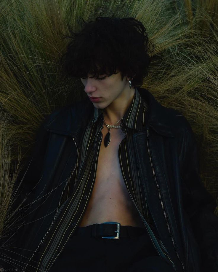 a man with no shirt sitting in the grass wearing a leather jacket and chain necklace