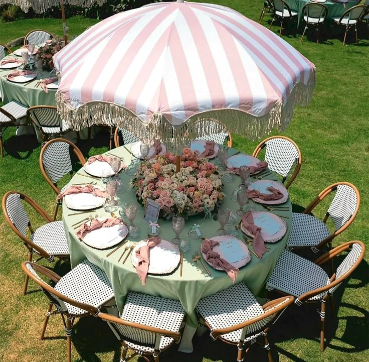 a table set up with plates and flowers under an umbrella in the grass at a wedding reception