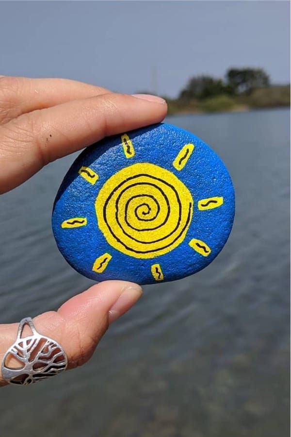 a hand holding a rock with a sun painted on it in front of the water