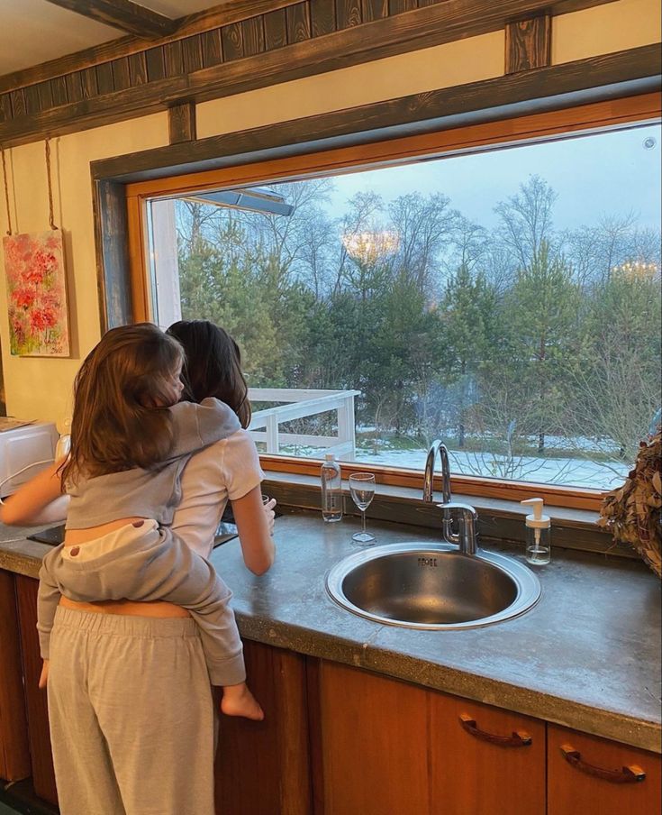 two children are standing in front of the kitchen window looking out at the snow outside