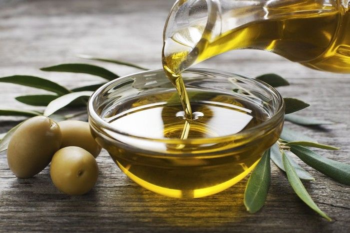 olive oil being poured into a glass bowl with two olives on the table next to it