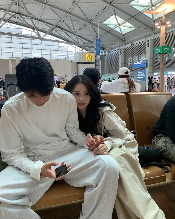 two people sitting next to each other on a bench at an airport, one holding a cell phone