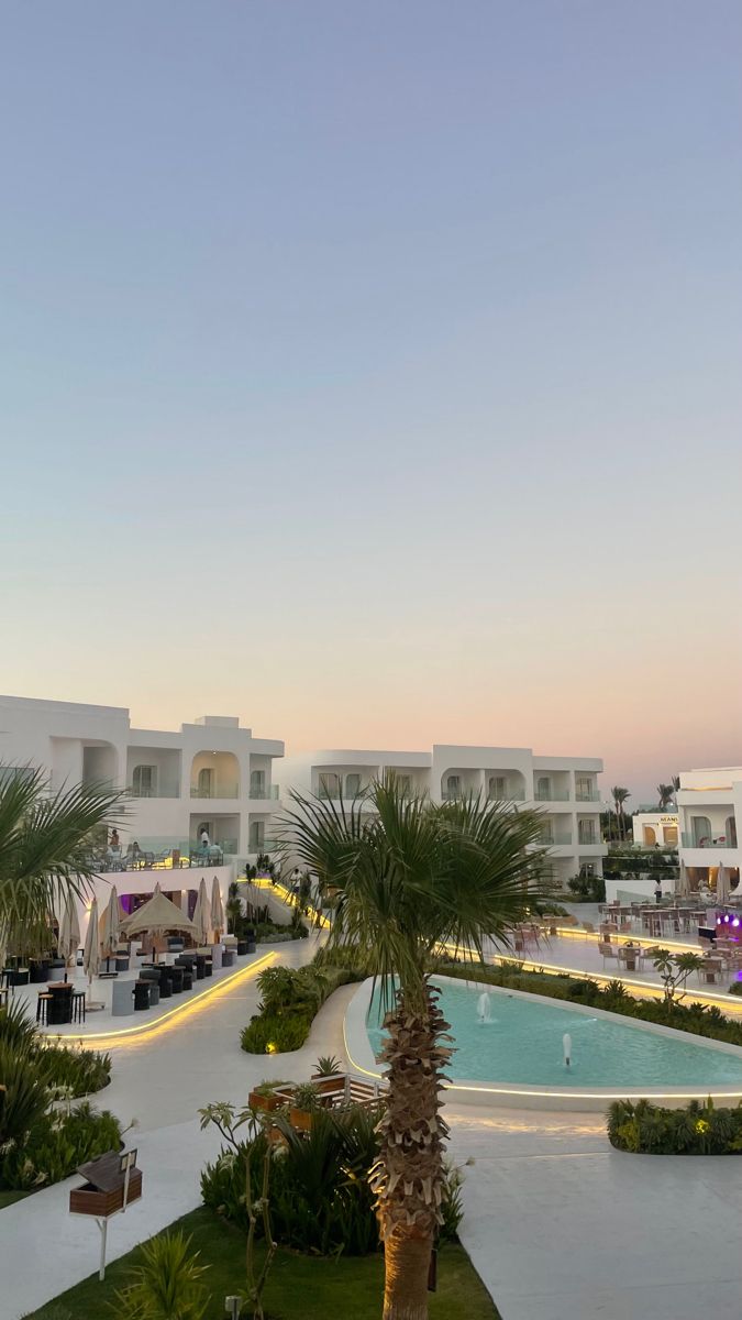 an outdoor swimming pool surrounded by palm trees and lawn chairs at dusk with buildings in the background