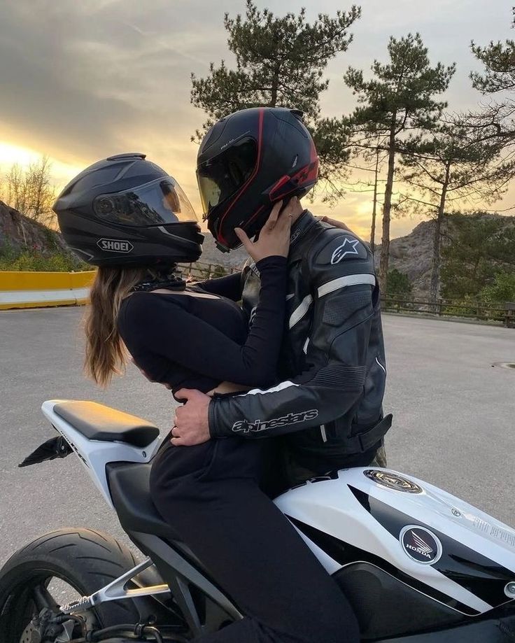 a man and woman sitting on a motorcycle talking on their cell phones while the sun is setting