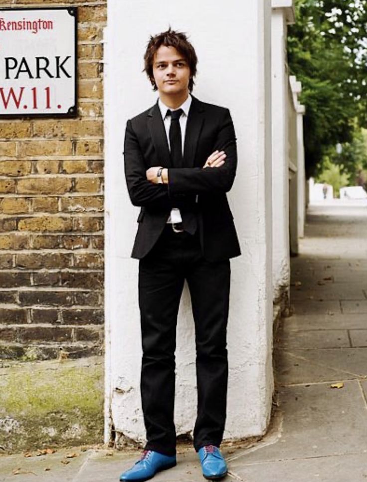 a man in a suit and tie leaning against a wall with his arms crossed, wearing blue shoes