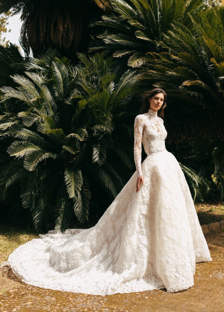 a woman in a white wedding dress standing next to some trees and palm trees with her hands on her hips