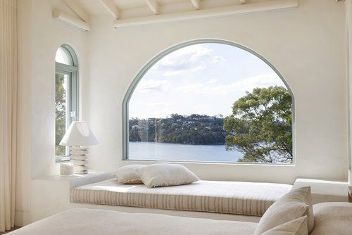 an arched window overlooks the water and trees from a bedroom with white bedding