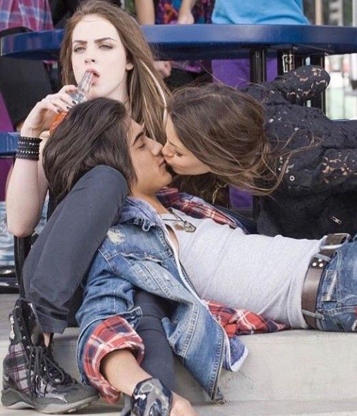 three young women sitting on the ground and kissing each other with people standing in the background