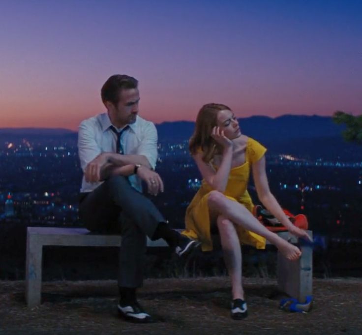 a man and woman sitting on a bench in front of a city at night with the sun setting