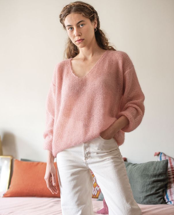 a woman standing in front of a bed wearing white pants and a pink sweater with an open v - neck