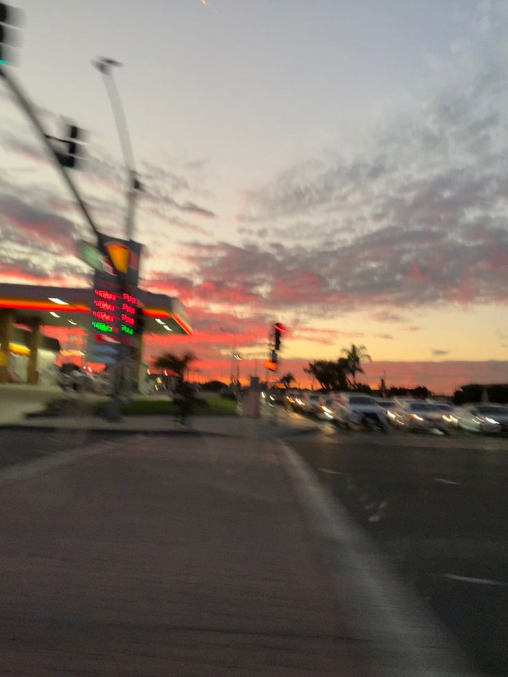 the sun is setting over a gas station and cars are driving down the street in front of it