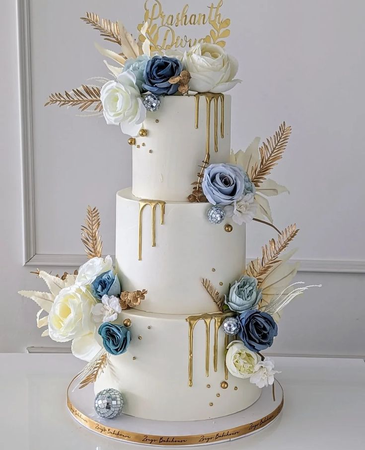 a three tiered wedding cake with blue and white flowers on the top, gold lettering