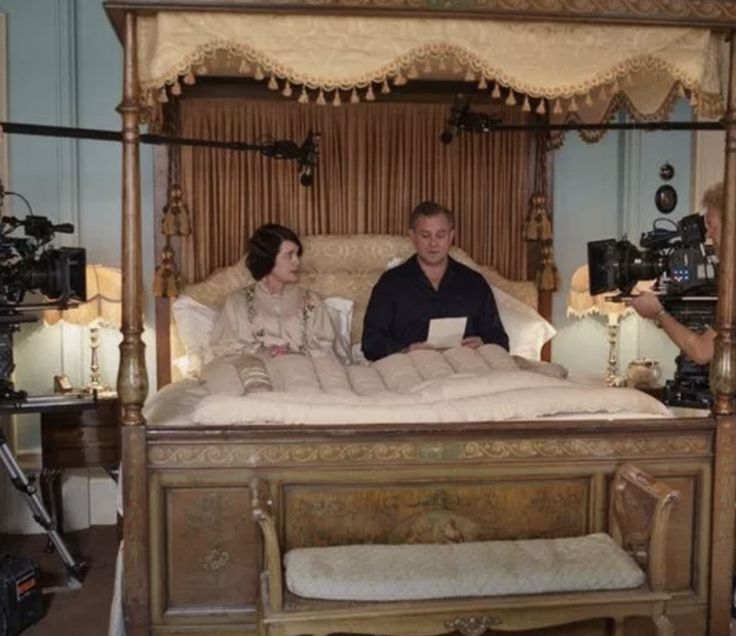 a man and woman sitting on top of a bed in a room with cameras around them