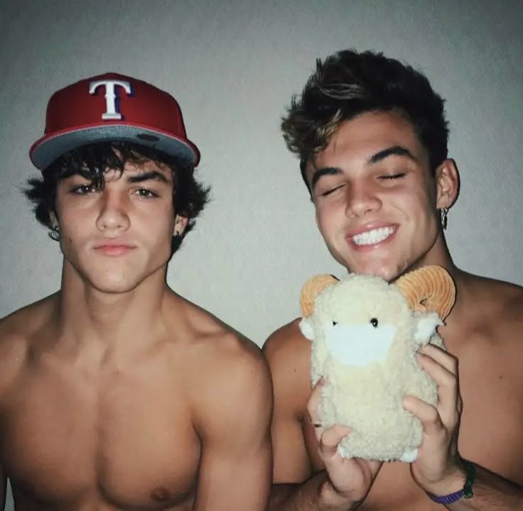 two young men are posing with one holding a stuffed animal and the other wearing a baseball cap