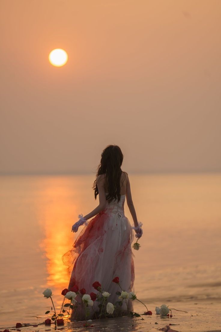 a woman in a dress standing on the beach looking at the water with flowers around her