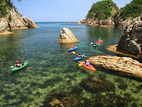 several people in kayaks paddling on the water near some rocks and trees, surrounded by clear blue water