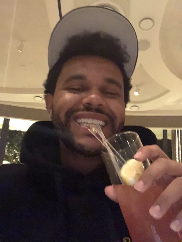 a man holding a drink in his right hand and smiling at the camera while wearing a hat