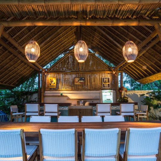 an outdoor dining area with white chairs and wooden tables in front of a bamboo roof