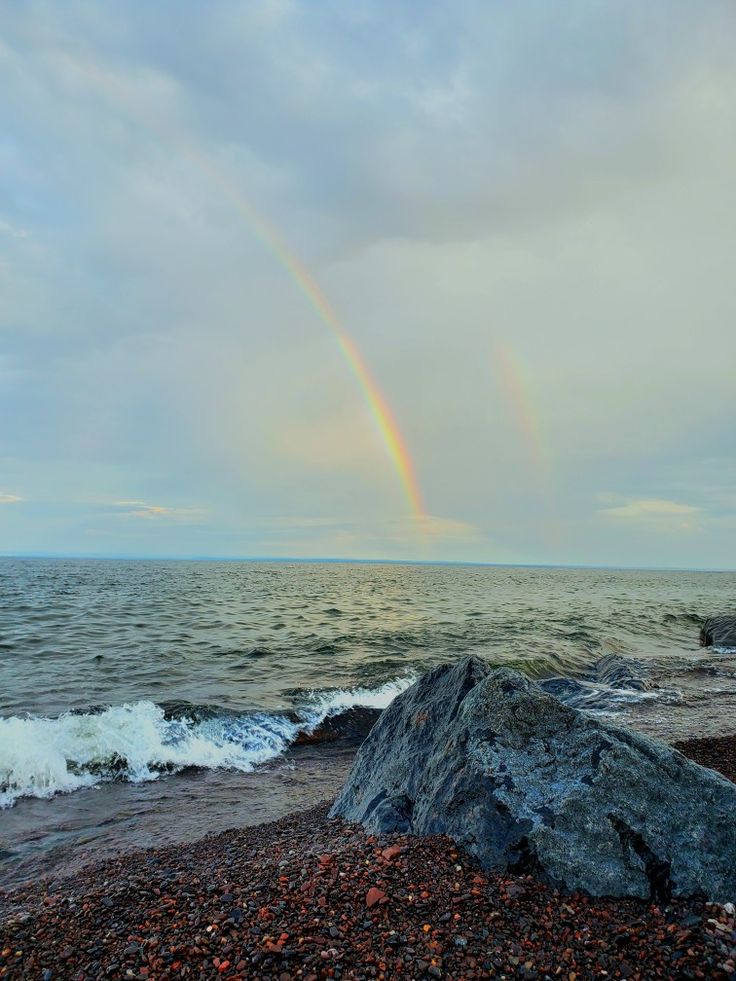 a rainbow shines in the sky over an ocean with rocks and pebbles on the shore