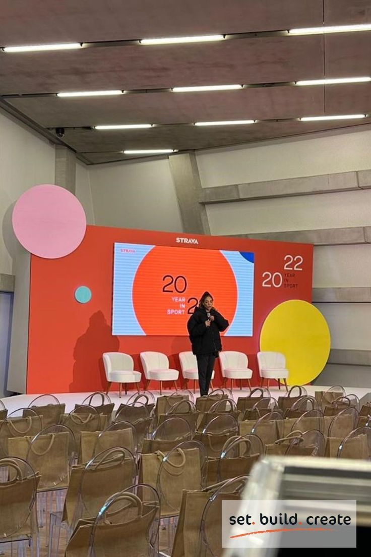 a woman standing in front of a large screen on a stage with lots of chairs